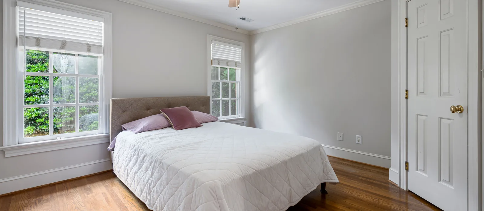 Sunlit bedroom with white quilted bedding, lavender pillows, large windows overlooking greenery, and warm hardwood floors